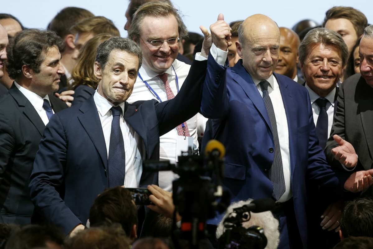 Vasemmalla seisova Nicolas Sarkozy ja oikealla seisova Alain Juppé pitävät toisiaan kädestä, käsi ylös kohotettuna. Taustalla pukuihin pukeutuneita miehiä.