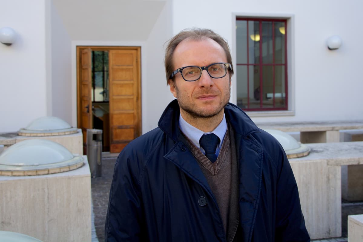 Silmälasipäinen dosentti Gino Scaccia sinisessä trenssitakissaan valkoisen rakennuksen luona.