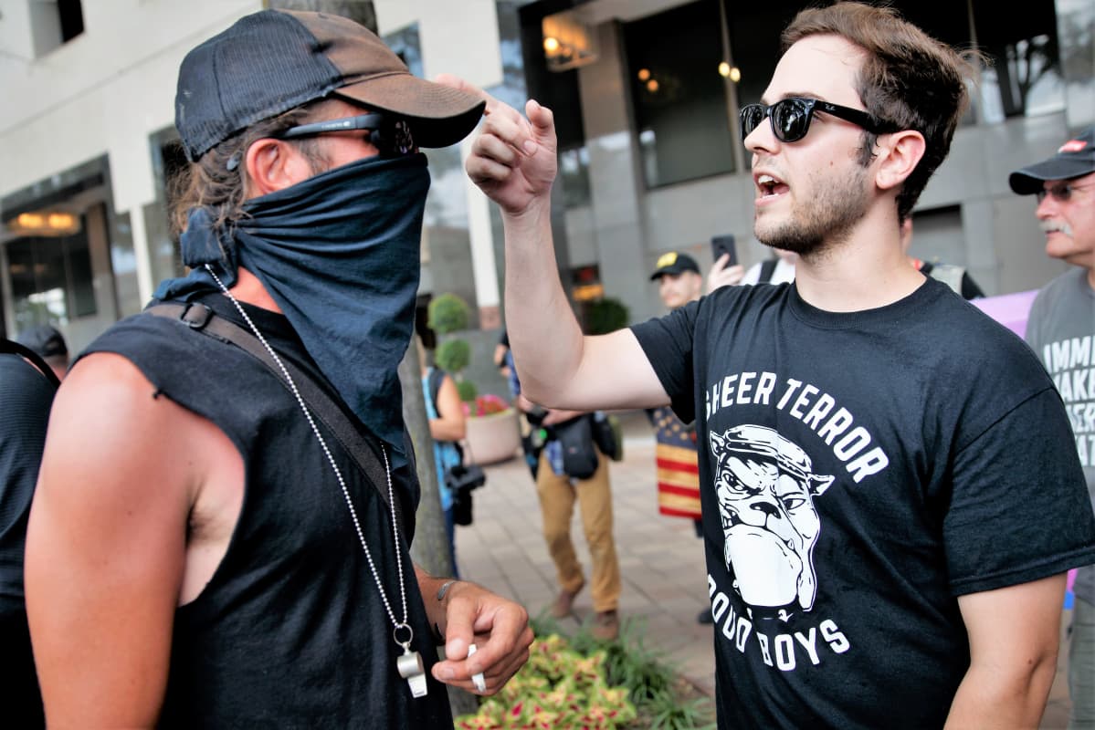 Antifan jäsen ja Proud Boysin jäsen keskustelevat kiivaasti. Antifan miehellä on musta hihaton paita, kaulassa narussa pilli, päässä lippalakki ja mustat lasit. Proud Boysien miehellä on mustat lasit ja t-paita, jossa on järjestön nimi ja bulldogin kuva.