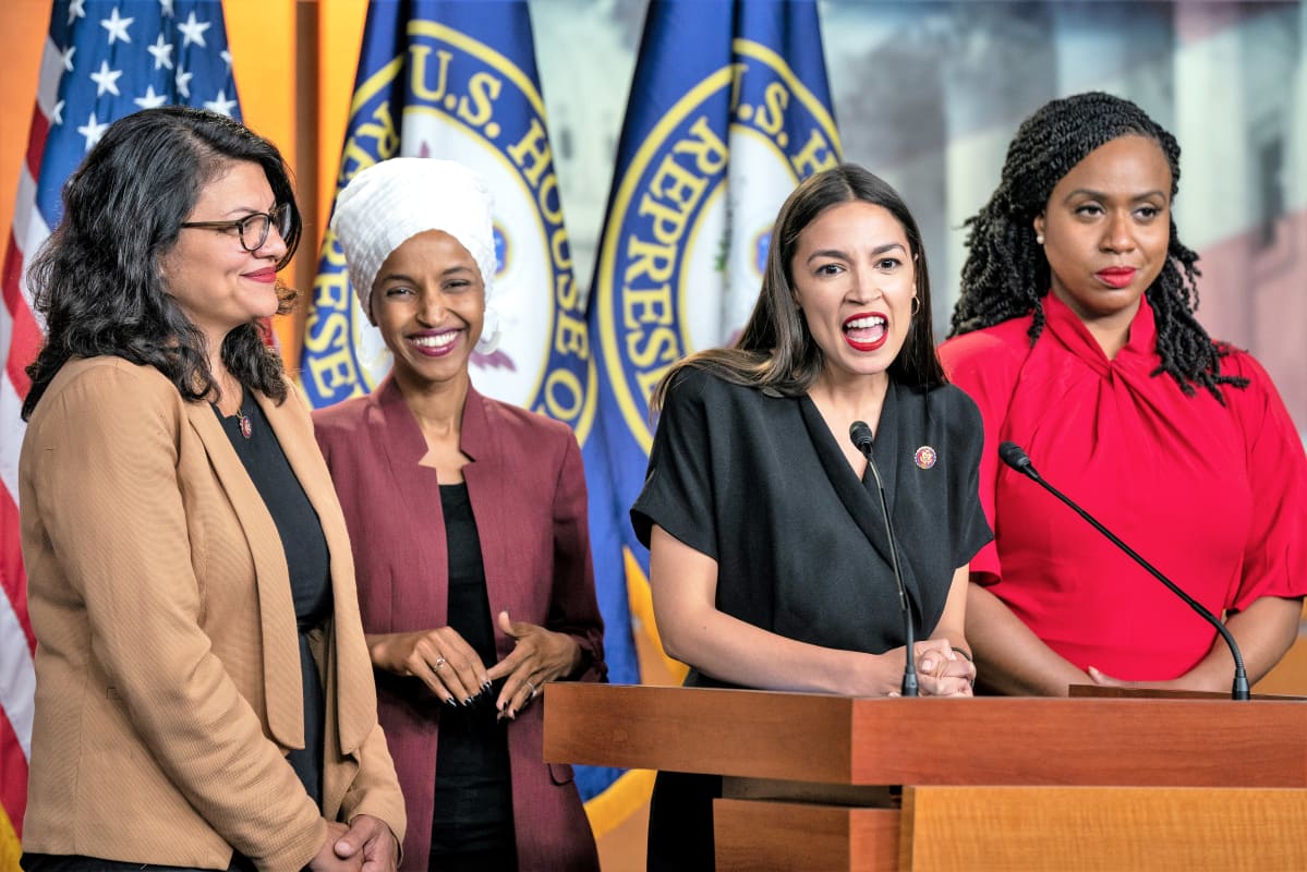 Neljä kongressiedustajaa seisoo Yhdysvaltain lipun ja edustajainhuoneen lipun edustalla. Alexandria Ocasio Cortez puhuu juuti mikrofoneihin.