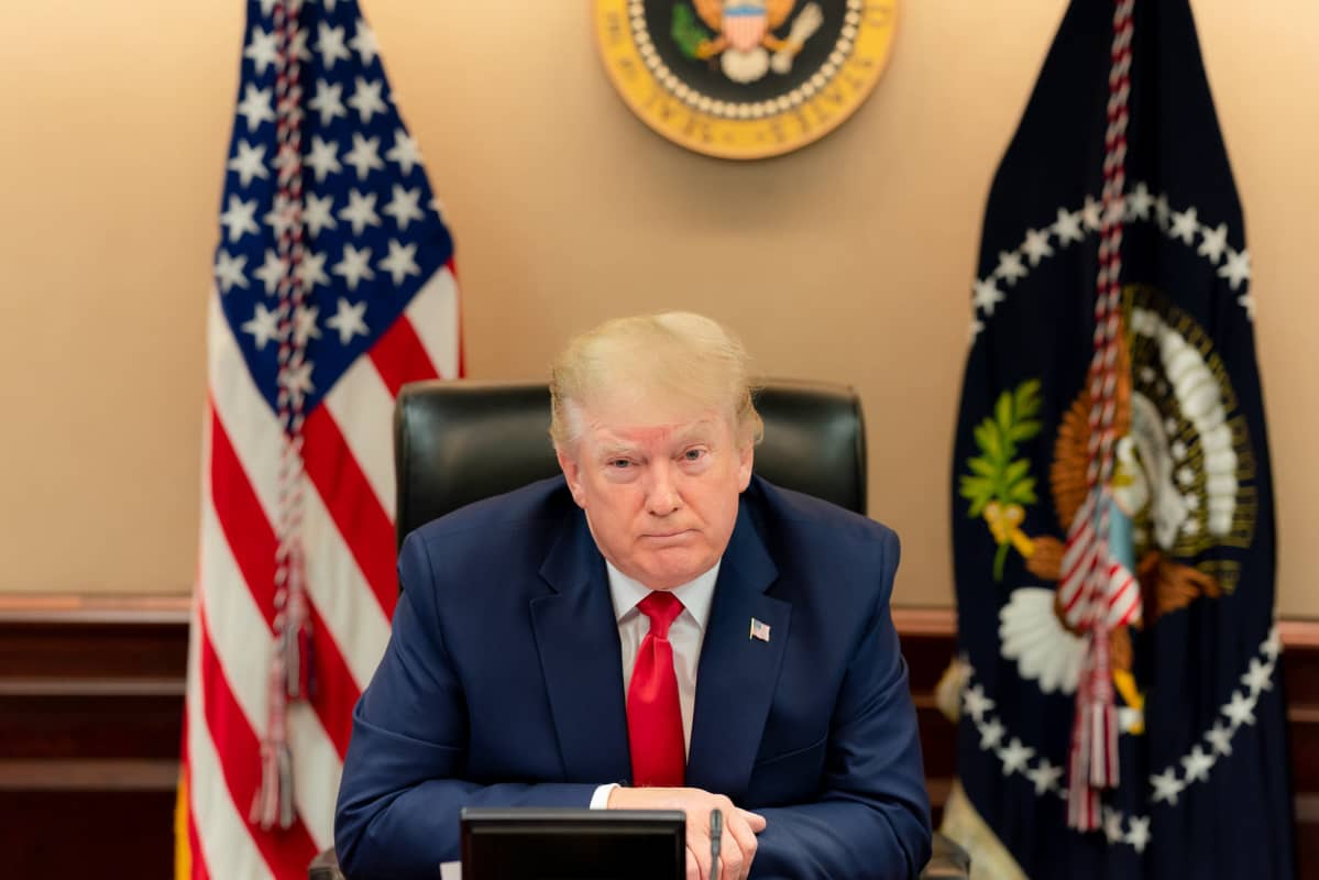 Donald Trump seuraa videoneuvottelua G20 kokouksessa. Taustalla USA:m ja USA:n presidentin liput.