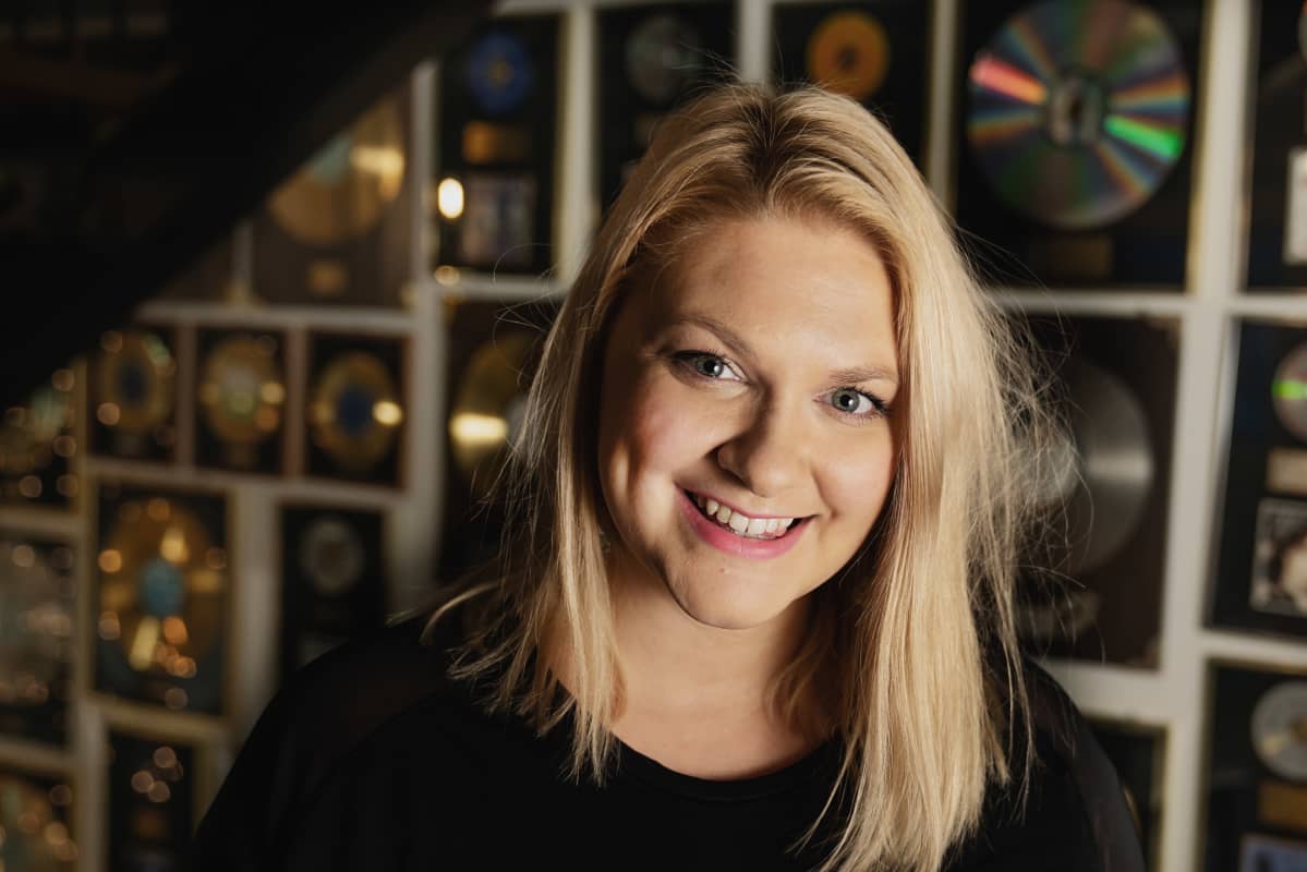 Karoliina Kanerva hymyilee lähikuvassa Warner Music Finlandin tiloissa, taustalla seinä täynnä kultalev.