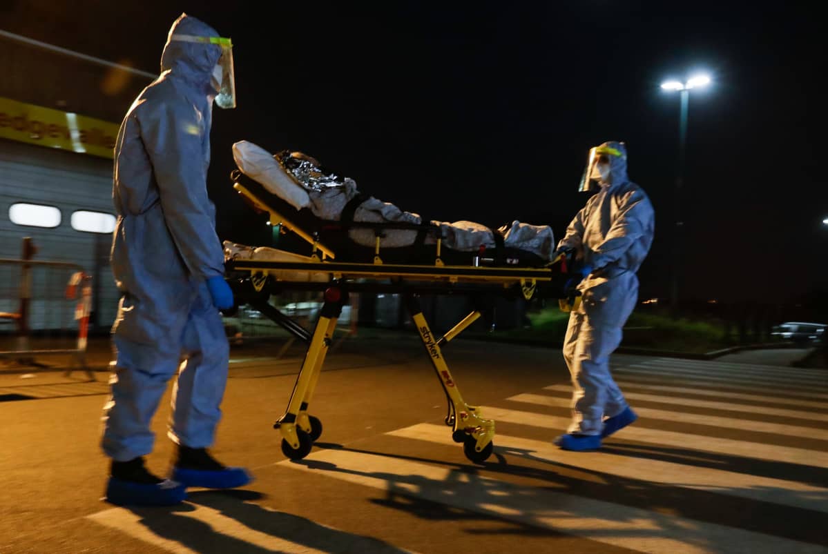 Två hälsovårdare rullar en sjuk äldre kvinna på en bår mot en ambulans. Det är natt och vårdare är klädda i heltäckande skyddsutrustning.