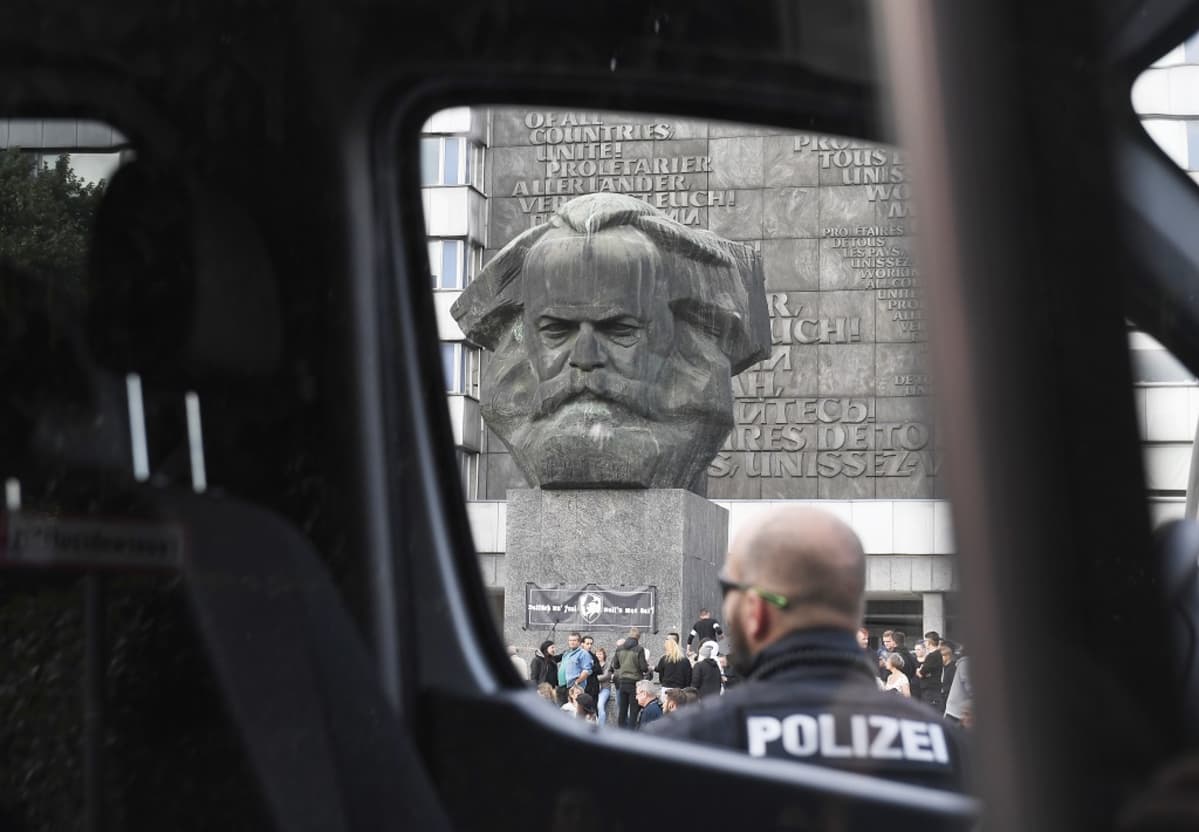 Poliisiauton ikkunan läpi otettu kuva. Etualalla auton luona seisoo kaljupäinen parrakas poliisimies. Aukiolla näkyy jylhän betoninen Karl Marxin pää.