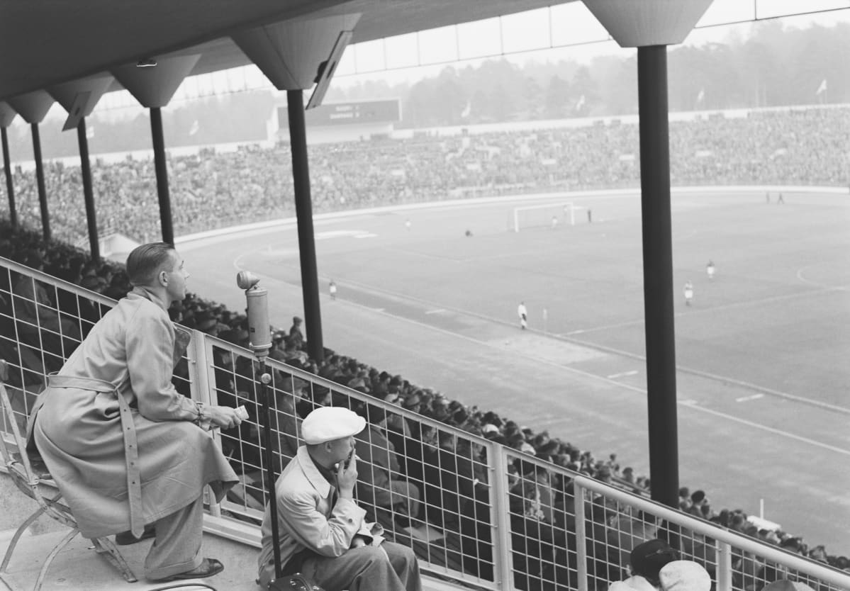 Helsingin olympiastadion. Jalkapallo-ottelu. Katsomo. Mikrofonin ääressä urheiluselostaja Enzio Sevón