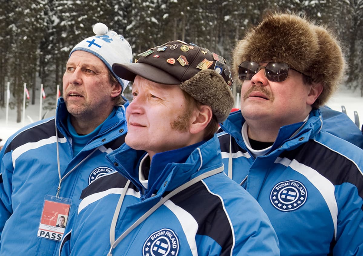 Suomen hiihtomaajoukkueen asuissa olevat miehet katsovat intensiivisesti urheilusuoritusta.