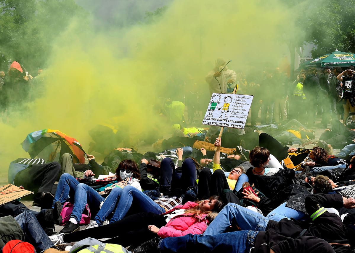 Keltaliivit ja ympäristöaktivistit protestoivat Bayerin & Monsanto- yhtiötä vastaan 18. toukokuuta. Protestoijat makaavat maassa, keltaista väripuuteria leijuu ilmassa ja yksi protestoija esittää ruiskuttavasna kemikaalia maassa olevien päälle.