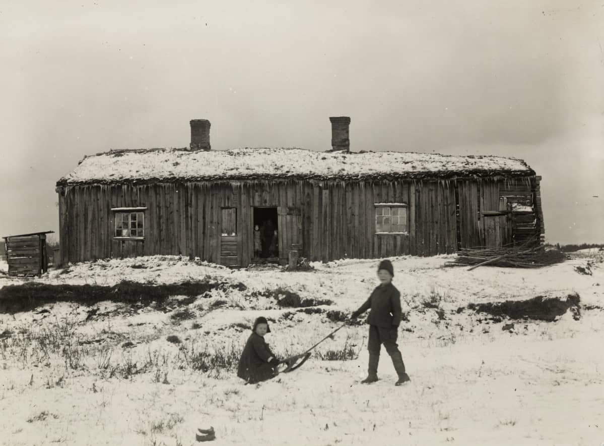 Vanha puinen rakennus, joka on rakennettu 1700-luvulla. Kuvan etualalla poika vetää toista poikaa kelkalla.