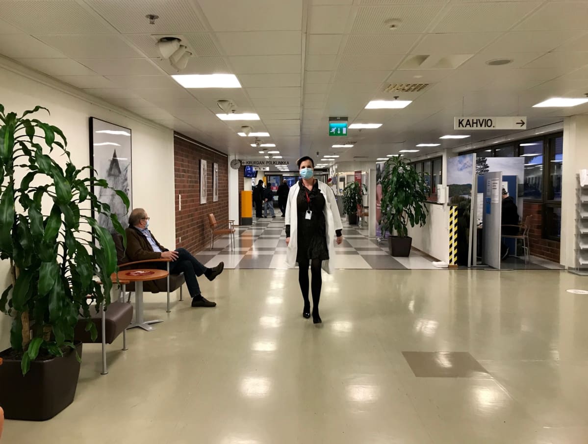 Lääkäri kävelee sairaalan aulassa maski naamassa, päällä lääkärintakki ja jalassa korkokengät.