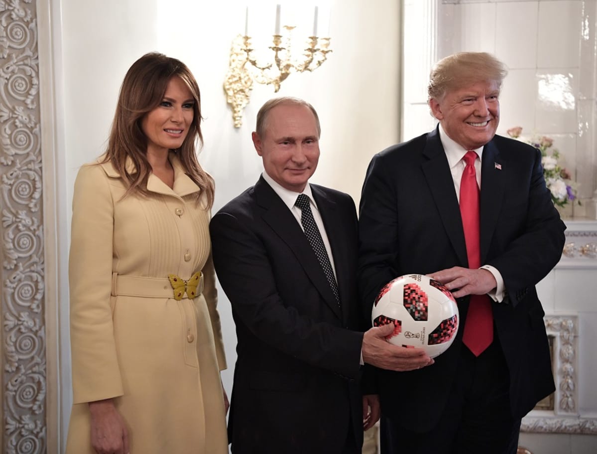 Melanialla on vaaleanruskea takki. Tummissa puvuissa olevat Trump ja Putin pitelevät jalkapalloa. Melania Trump ja Putin hymyilevät vienosti, Trump leveästi.