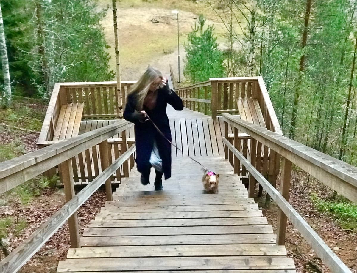 Vaalea, pitkätukkainen nainen lilassa pitkässä takissa juoksee ylös kuntoportaita pienen koiran kanssa