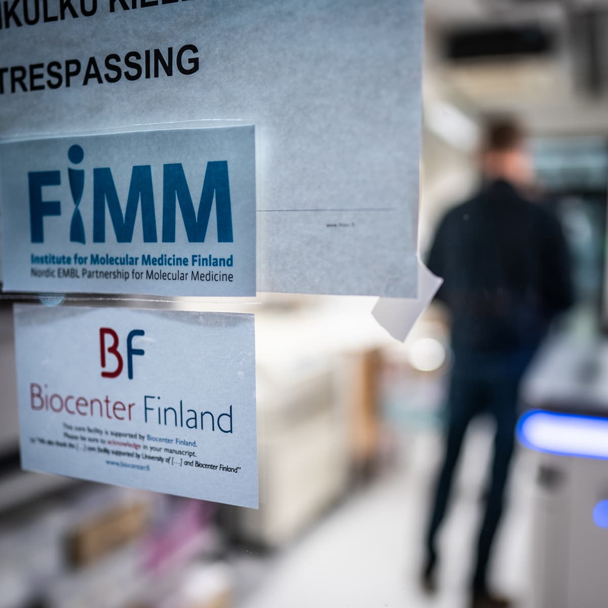 FIMM-teksti paperilapussa laboratorion lasiovessa, oven läpi näkyy mieshahmo.