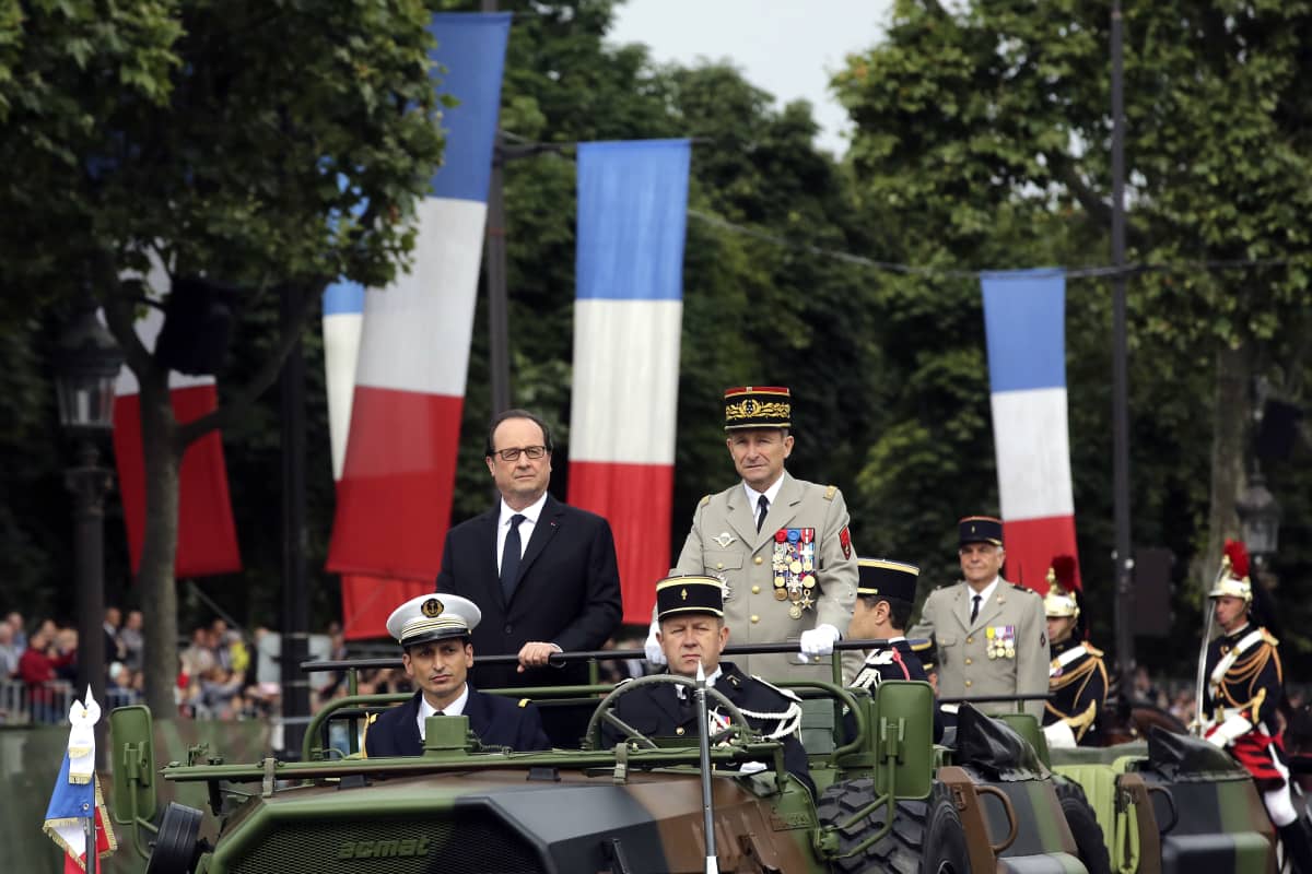 Ranskan presidentti François Hollande ja asevoimien komentaja Pierre de Villiers seisovat autossa, jota armeijan auton takaosassa. Saattueessa on muitakin sotilaiden ajamia autoja. Katua reunustavat puut j Ranskan liput.