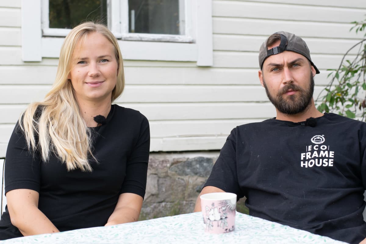 Ett ca 30-årigt par, en man och en kvinna, sitter utomhus vid ett trädgårdbord. De ser mot kameran. I bakgrunden ett vitt trähus.