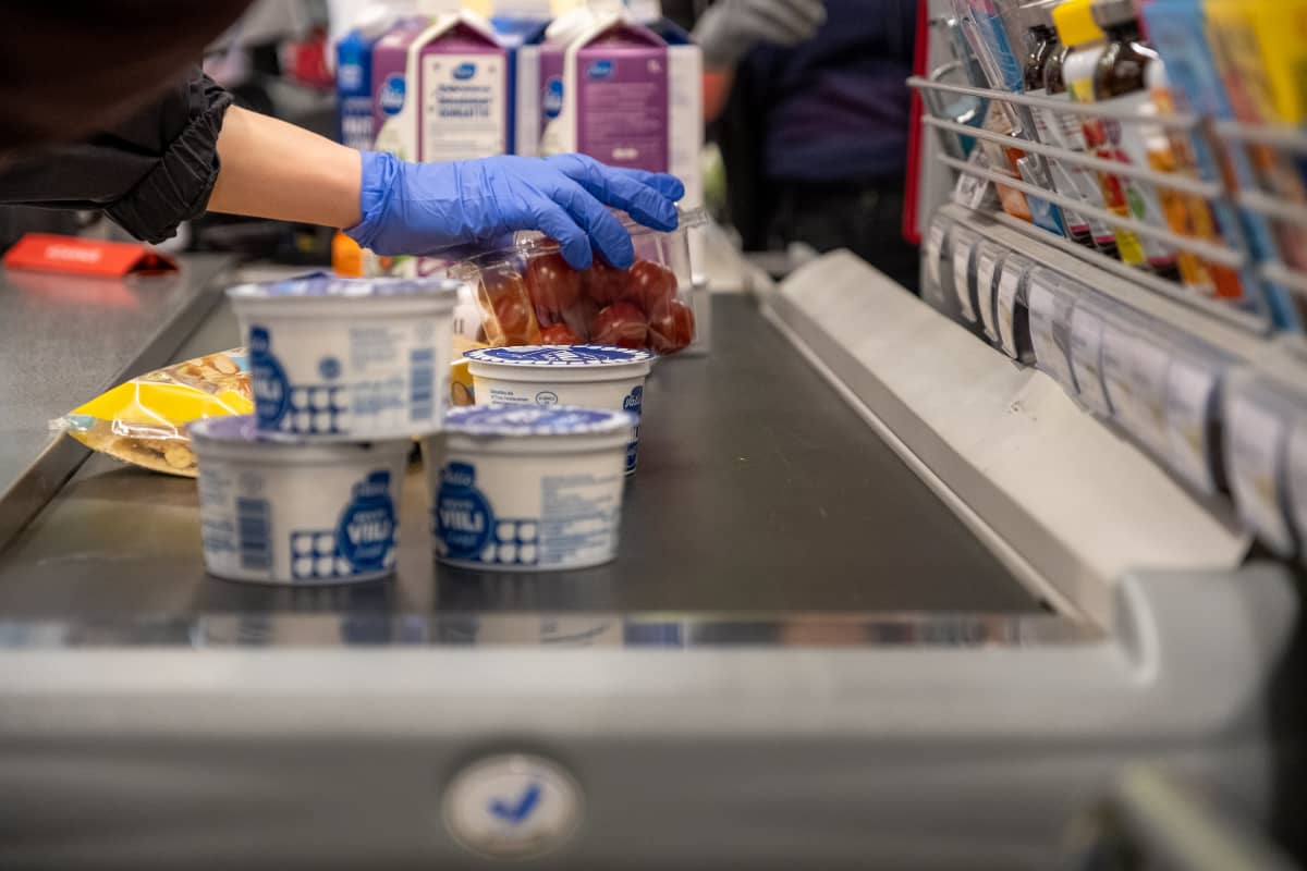 Suojakäsineellä suojattu käsi nostaa ostoksia elintarvikemyymälän liukuhihnalle.