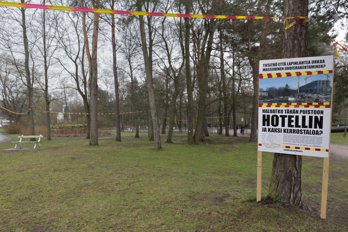Kyltti, jossa lukee Haluatko tähän poistoon hotellin ja kaksi kerrostaloa", ja muovinauhalla merkattu rakennuksen paikka Lapinlahden sairaalan puistossa.