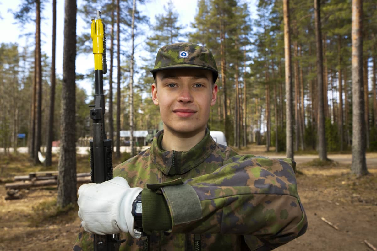 Varusmiessoittokuntaan kuuluva Lauri Heroja pitelee kivääriään ja katsoo kameraan metsässä.