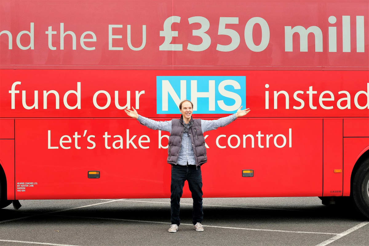 Dominic Cummingsia esittävä Benedict Cumberbatch seisoo kädet levällään punaisen kaksikerroksisen bussin edessä. Bussin kyljessä on brexit-kampanjan iskulauseita.