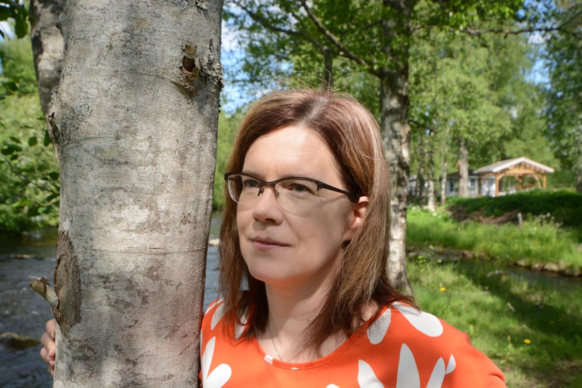 Sari Hyttinen nojaa puuhun ja katselee joelle.