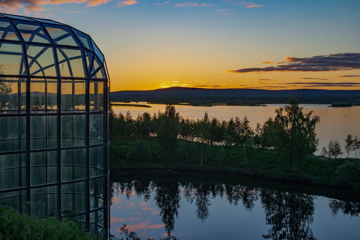 Keskiyön aurinko laskee Ounasjoen taakse, etualalla Arktikum-talon lasikattoa.
