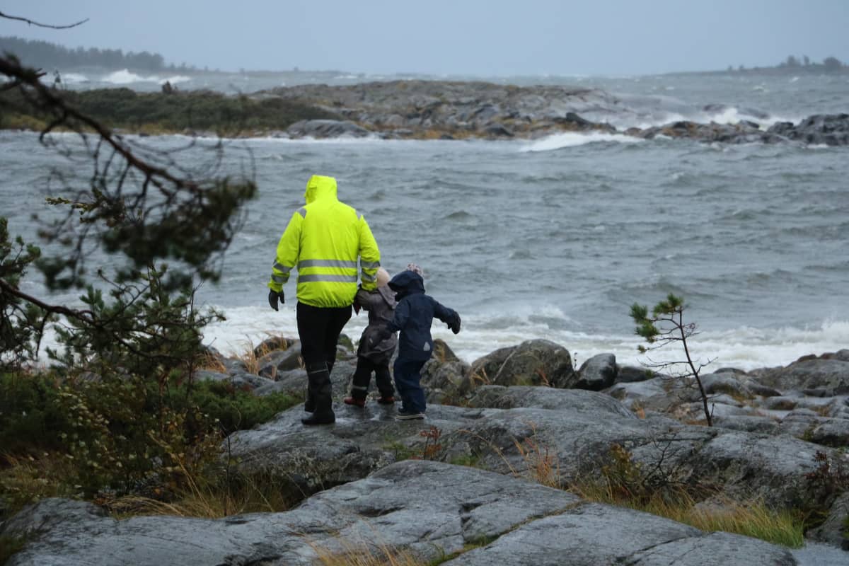 En pappa med två barn går på klippor i hård vind. Vattnet slår mot klipporna.