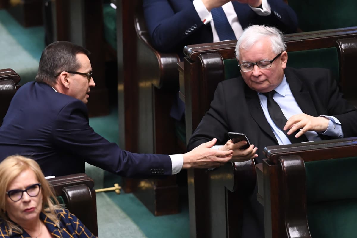 Mateusz Morawiecki näyttää kännykkän näyttöä Jaroslaw Kaczynskille parlamenttisalin käytävän yli