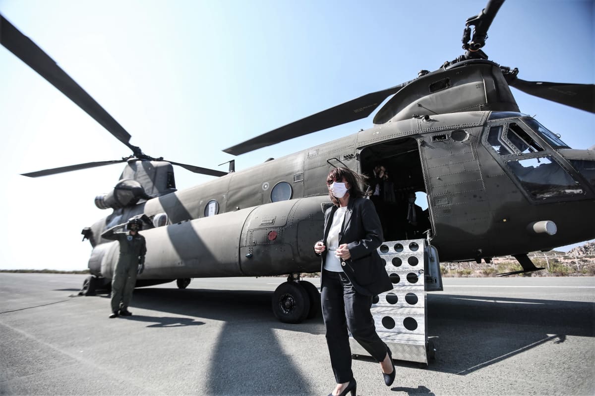 Kreikan presidentti Katerína Sakellaropoúlou on tulossa helikopterista. Hänellä on harmaa asu. Taustalla lentäjänasuinen kypäräpäinen mies vetää kättä lippaan presidentille.