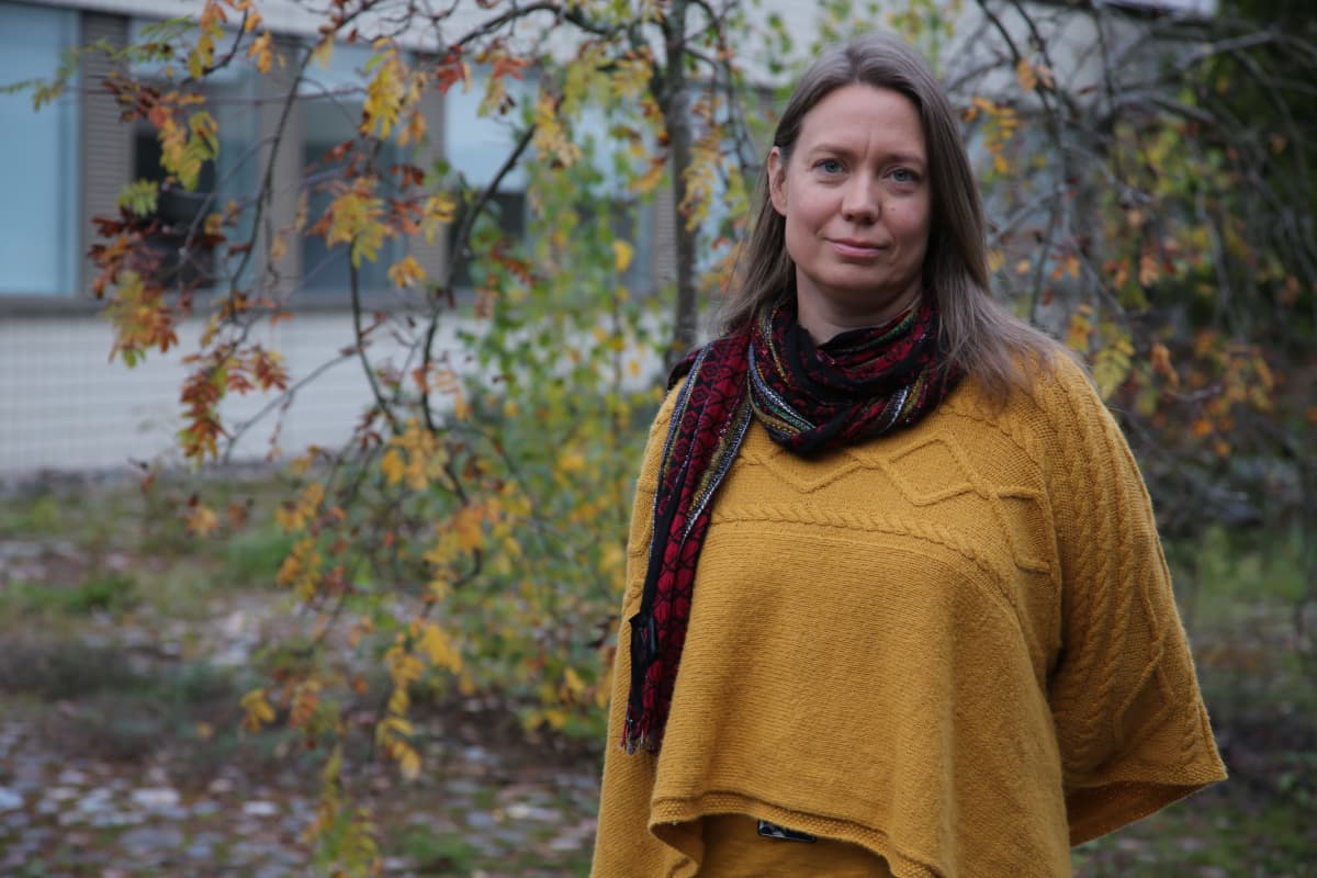 Tummahiuksinen nainen seisoo puutarhassa päällään keltainen villatakki ja punainen kaulahuivi.
