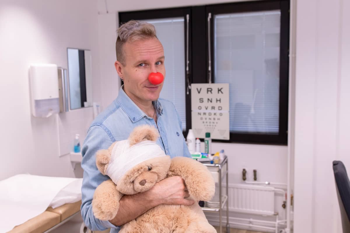 Puoli seitsemän -ohjelman juontaja Mikko Kekäläinen lääkärissä nalle sylissään, Mikolla on punainen Nenäpäivä-nenä.