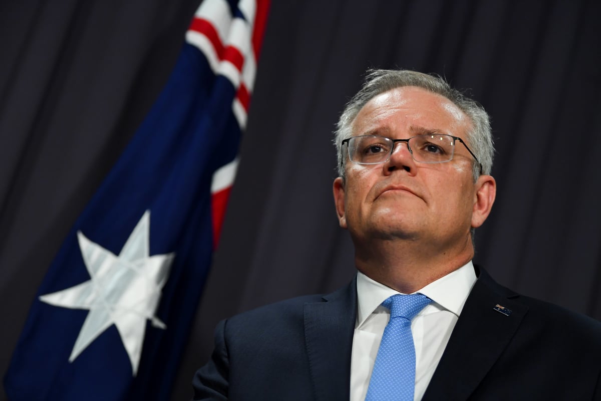 Scott Morrison on mustan taustakankaan edessä. Hänellä on tumma puku ja vaaleansininen kravatti. Taustalla näkyy Australian lippu.
