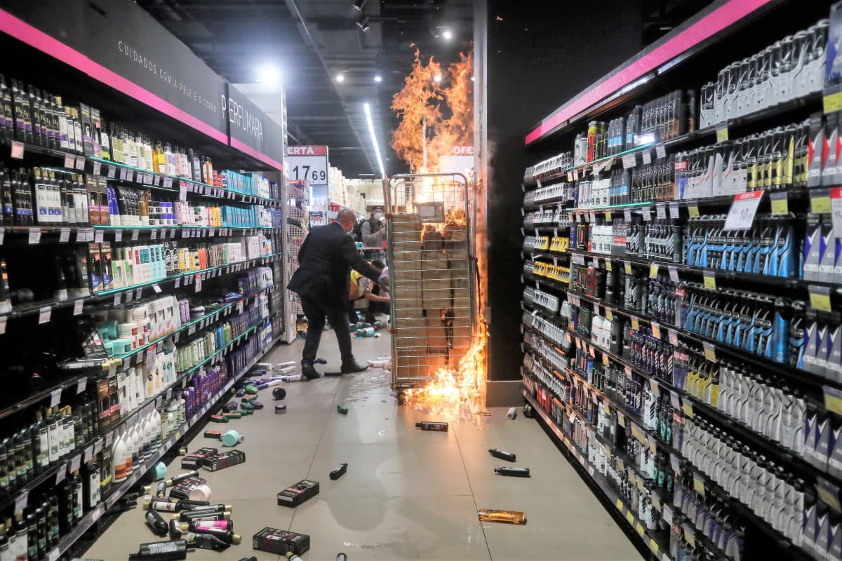 Ostoskorit palavat supermarketin käytävällä. Hyllyjen välissä lattialla on erilaisia pulloja ja astioita. Tummapukuinen mies on kulkemassa palavien ostoskorien ohi.