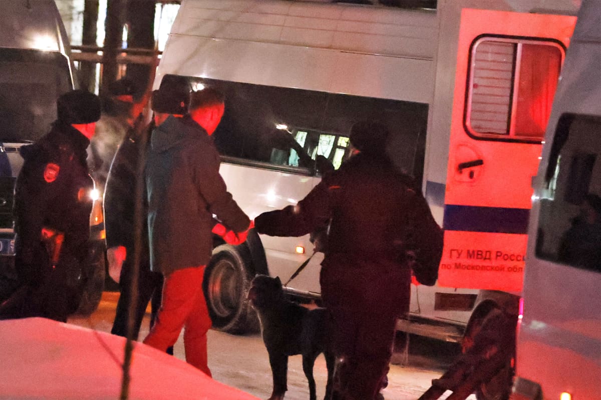 Himkin pöliisiaseman pihamaa. Kolme poliisi ohjaa Aleksei Navalnyia pakettiautoon. Auton takavalot värjäävät maiseman punaiseksi. Yhdellä poliisilla on koira hihnan päässä.
