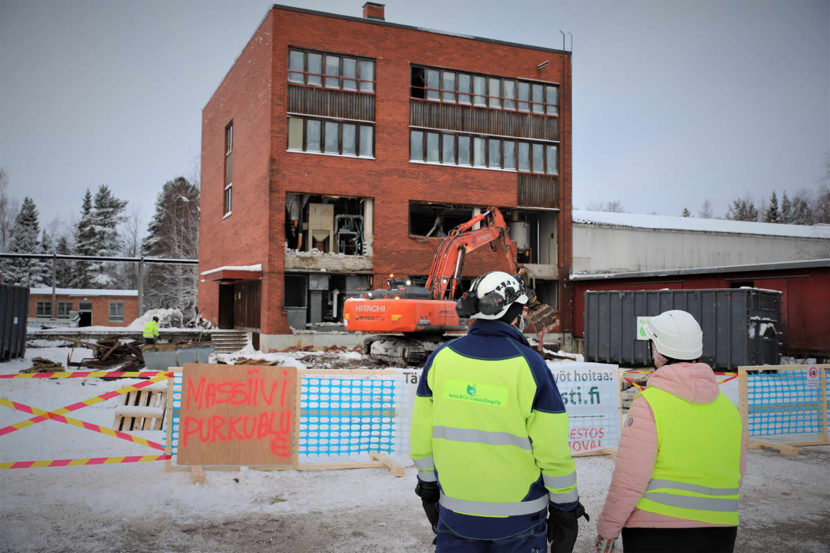 Kaivinkone hajottaa tiiliseinää, kaksi henkilöä tarkkailee purkua