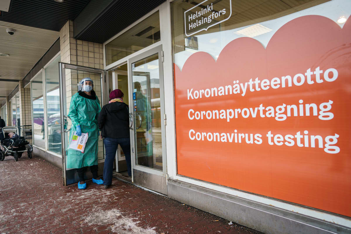Henkilö menossa sisään koronatestauspaikkaan. Henkilökunnan edustaja pitää ovea auki. Ikkunassa teksti koronanäytteenotto kolmella kielellä.