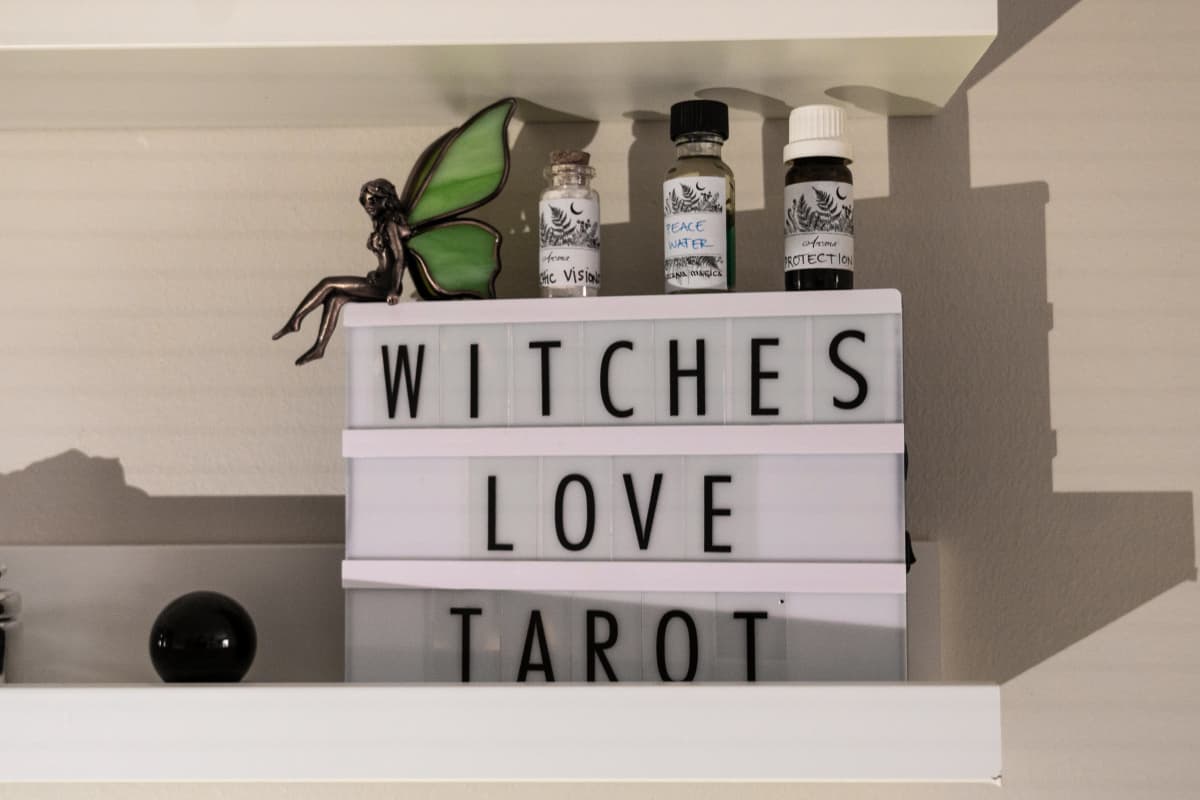 Neliön muotoinen pöytävalo, jossa on teksti "witches love Tarot". Lampun päällä on öljyjä ja keijukoriste.