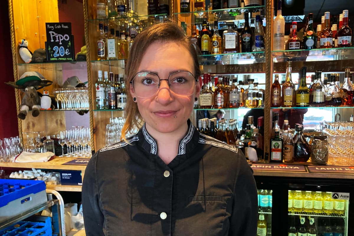 Silmälasipäinen nainen mustassa ravintolakokin takissa seisoo ravintolan tiskin takana, taustalla peilejä ja viinapulloja.