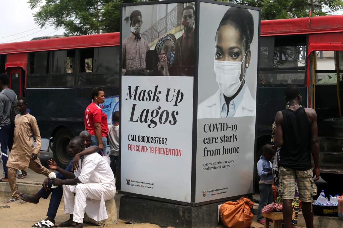 Ihmisiä istui bussipysäkillä Lagosissa kyltin edessä, jossa luki "mask up Lagos".