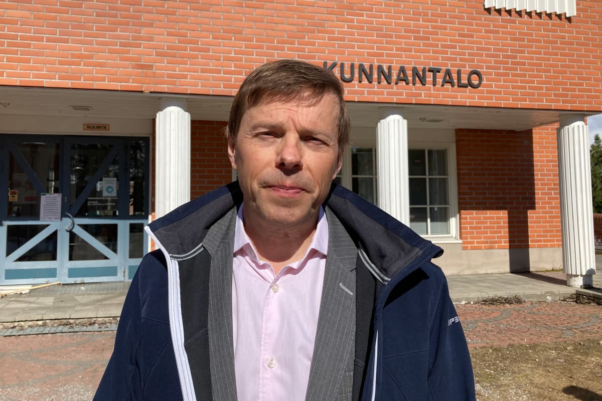 Timo Kurula katsoo kameraan silmiään siristellen ja takki auki, taustallaan Kittilän kunnantalo.