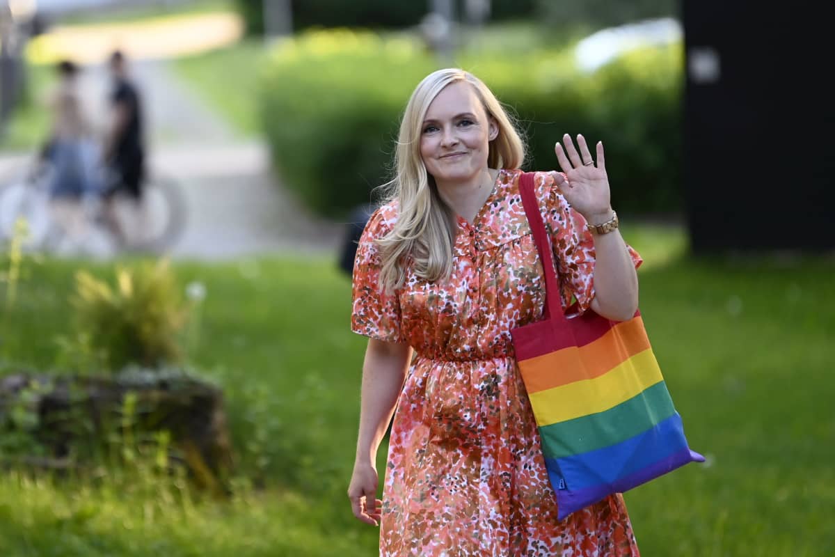 De Grönas ordförande Maria Ohisalo fotad ute. Hon har en röd klänning på sig och en tygkasse med Pride-färger. Hon vinkar mot kameran.