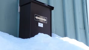 Kuvassa puutalon seinässä oleva postilaatikko. Lumikinos yltää laatikon alareunaan asti.