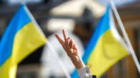 Henkilö näyttää voitonmerkkiä sormillaan Ukrainan lippuja taustalla.