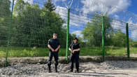 Photo shows two border guards standing in front of a section of fencing, which is about 4 metres tall.