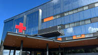 Ingången till sjukhus, glasväggar och ett stort rött kors på taket vid ingången.