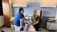 Matti Mikkola makaa sairaalasängyllä. Kaksi hoitajaa auttaa miestä sänkyyn.