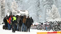 Joukko ihmisiä seisoo jonossa autopuomin edessä. Vieressä rivissä polkupyöriä lumisessa maisemassa. Rajavartija kävelee jonon vierellä.