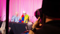 Windows95man and Henri Piispanen both dressed in colourful clothing, sitting a table at a press conference with a pink curtain in the background.