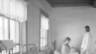Ikkunoiden vieressä neljä sänkyä, jossa kahdessa makaa lapsia ja perimmäisessä sängyssä äiti istuu vauva sylissään. Äidin vieressä seisoo nuori tyttö.
