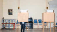 Kaksi puista äänestyskoppia suuressa tilassa, toisessa äänestyskopissa on henkilö, takaseinällä tuoleja siisteissä pinoissa, musta piano, sekä muotokuva vanhasta miehestä.