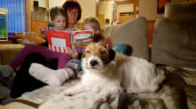 Penttilän perhe, äiti Liisa, Jade ja Helmi istuvat sohvalla ja lukevat kuvakirjaa. Perheen koira makaa vieressä.