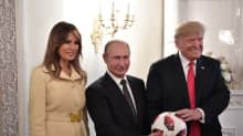 Melanialla on vaaleanruskea takki. Tummissa puvuissa olevat Trump ja Putin pitelevät jalkapalloa. Melania Trump ja Putin hymyilevät vienosti, Trump leveästi.